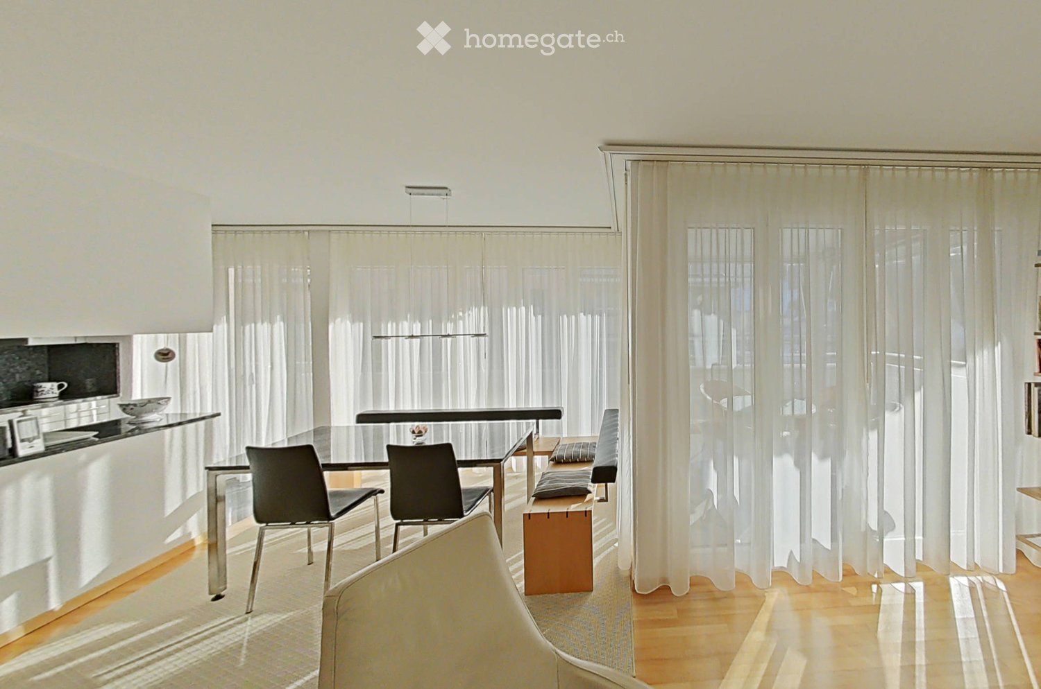Bright, modern living and dining area with large windows covered by sheer curtains, a sleek black dining table with chairs, a wooden bench, and a white sofa. The kitchen area features a black countertop with a cup and bowl.