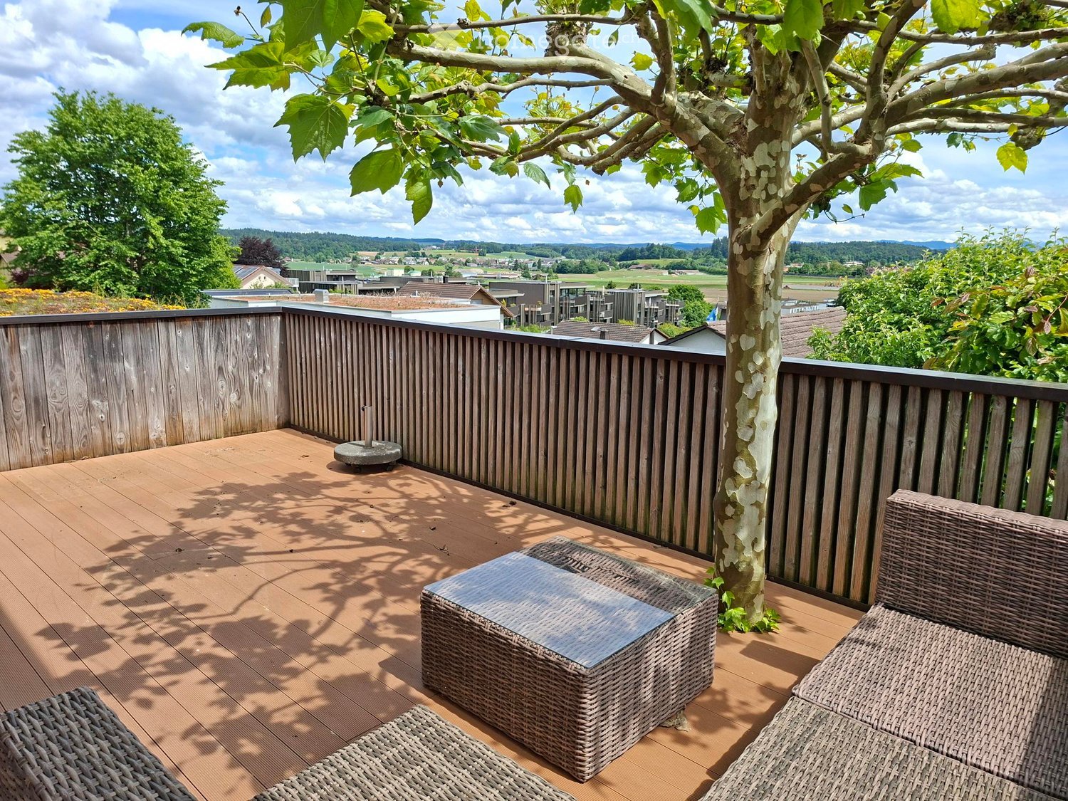 Balcony / Terrace. Wooden deck with wicker furniture, including a sofa, table, and chairs, surrounded by a wooden railing and a tree, with a scenic countryside view.