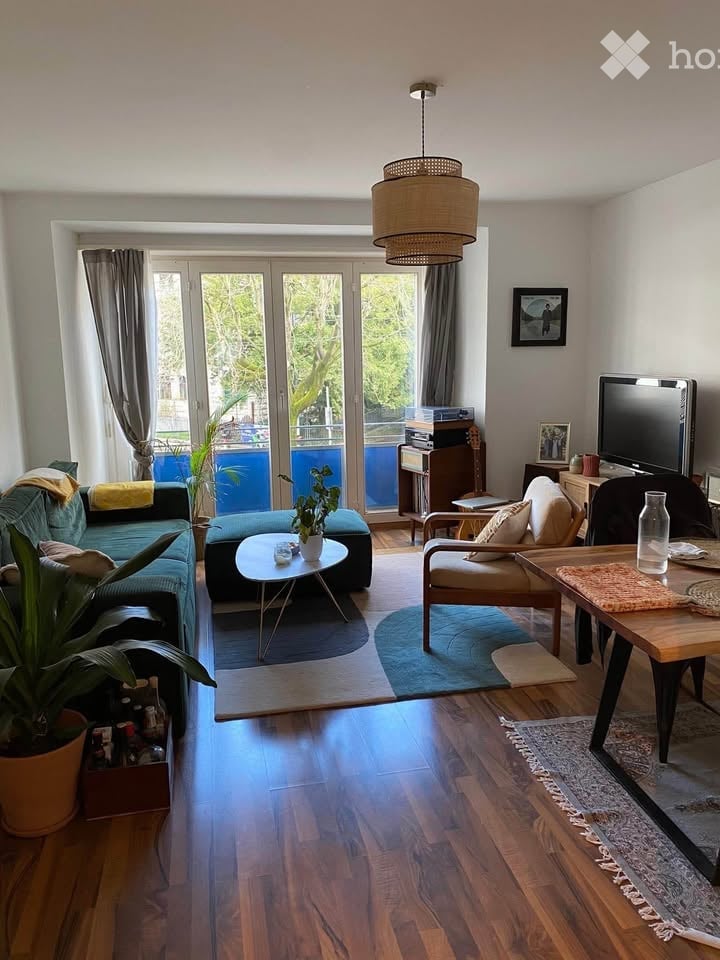 Modern living room with wooden flooring, large sliding glass doors leading to a balcony, a sofa, coffee table, armchair, dining table, television, and decorative plants.