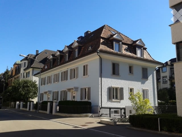 A multi-story residential building with a classic European architectural style, featuring white walls, dark brown roof, multiple windows with shutters, and a small garden area in front.