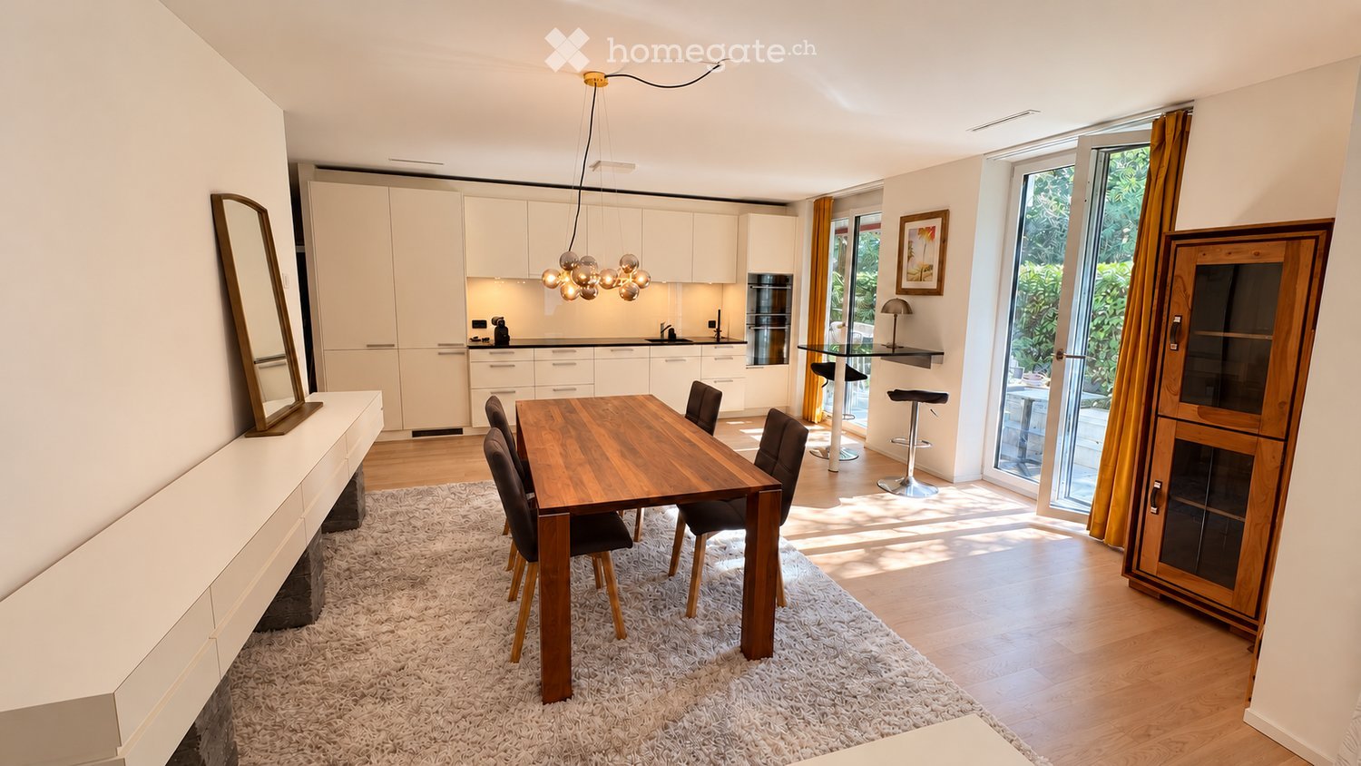 Modern open-plan dining and kitchen area with a wooden dining table, six chairs, white kitchen cabinets, a sink, and a bar area with stools. The space features a large mirror, a picture frame, and a hanging light fixture.