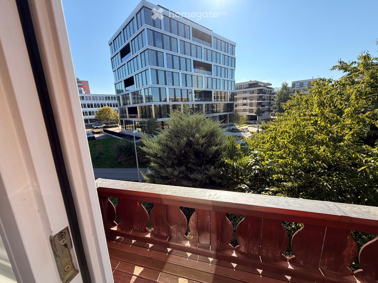 Balcony with wooden railing overlooking a modern urban area with tall buildings, greenery, and parked cars.