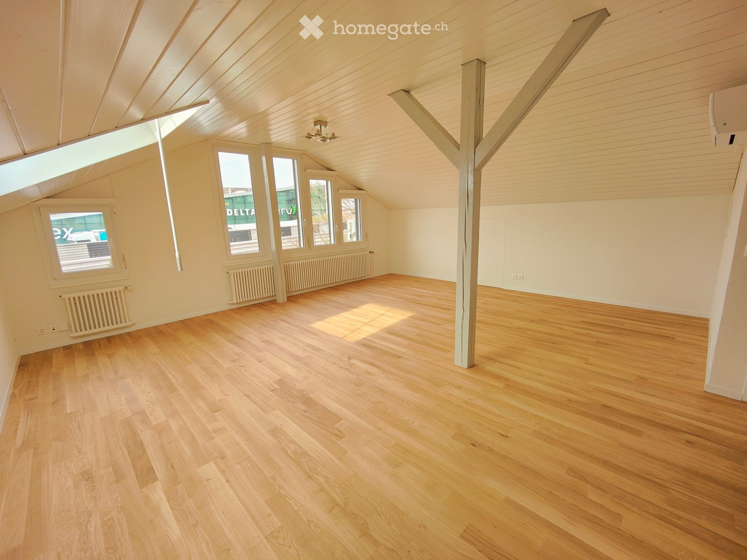 Empty attic room with wooden flooring, exposed wooden beams, and large windows allowing natural light. Radiators are visible along the walls.