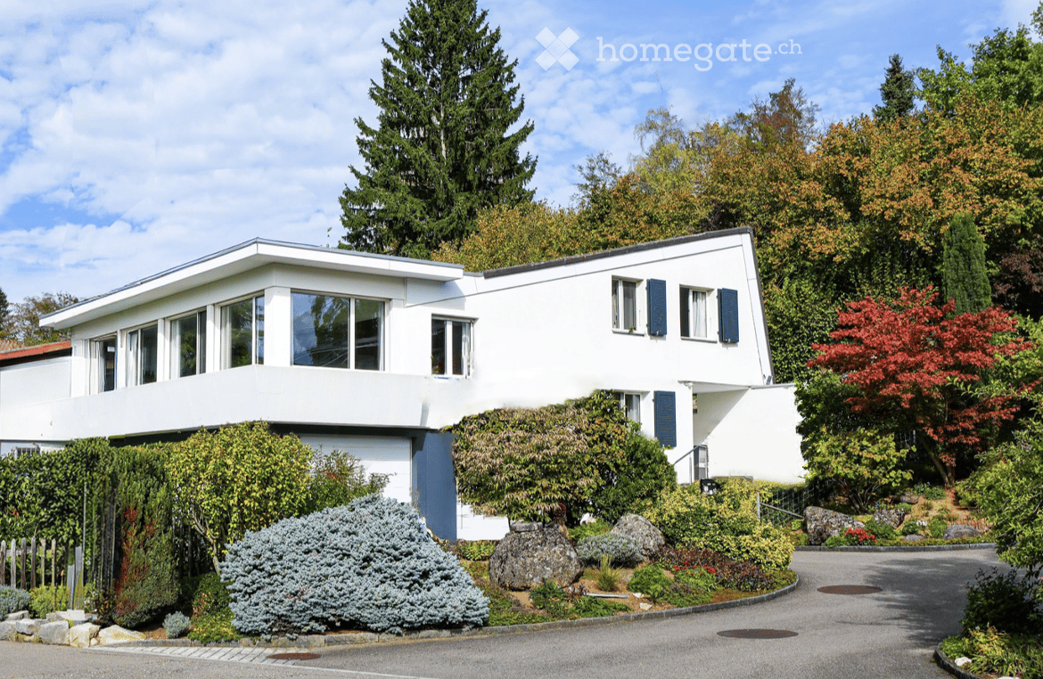 Modern two-story white house with large windows and blue shutters, surrounded by lush greenery and a well-maintained garden.