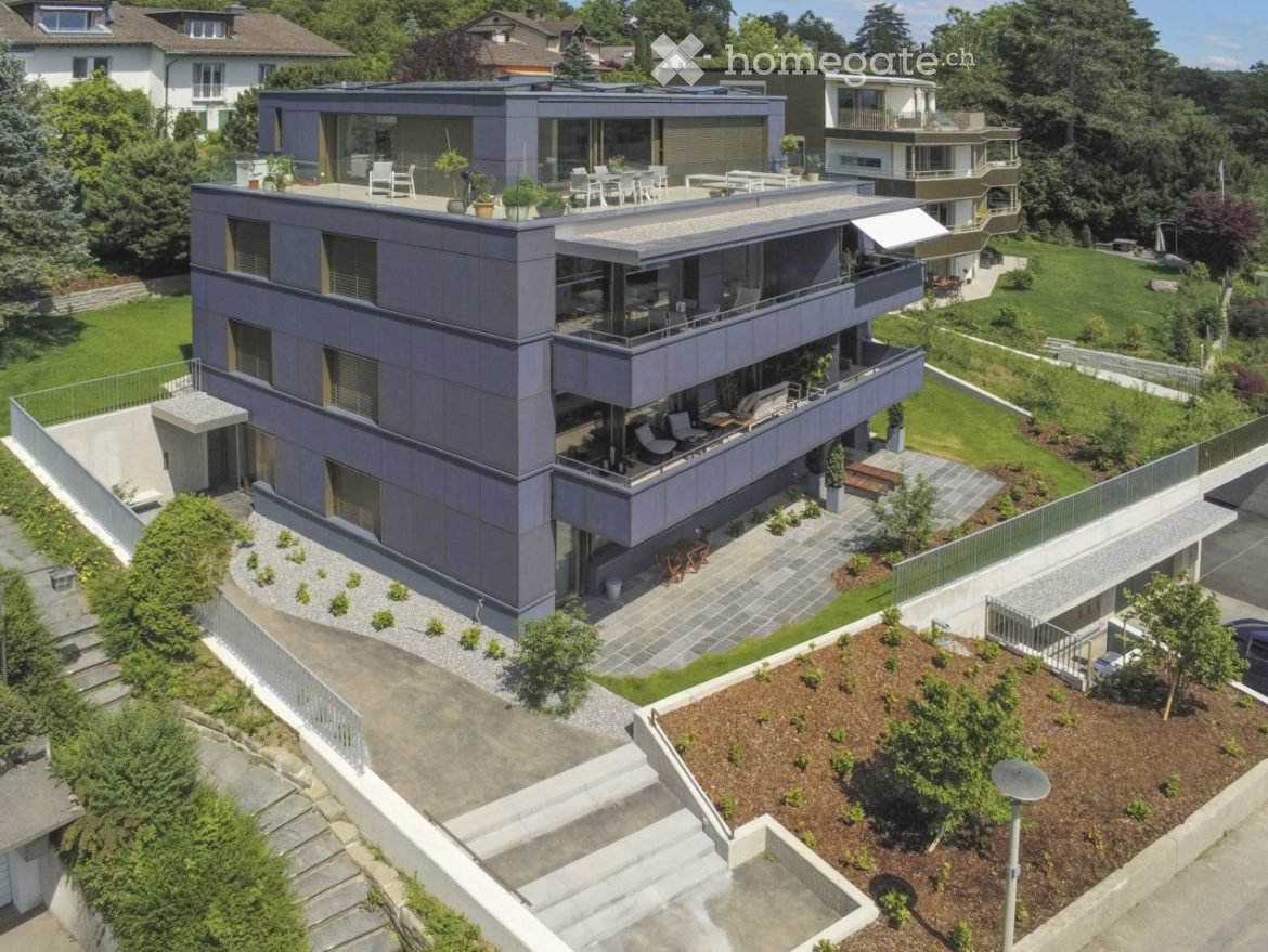 Modern two-story house with grey exterior, large windows, balconies, and surrounding greenery.