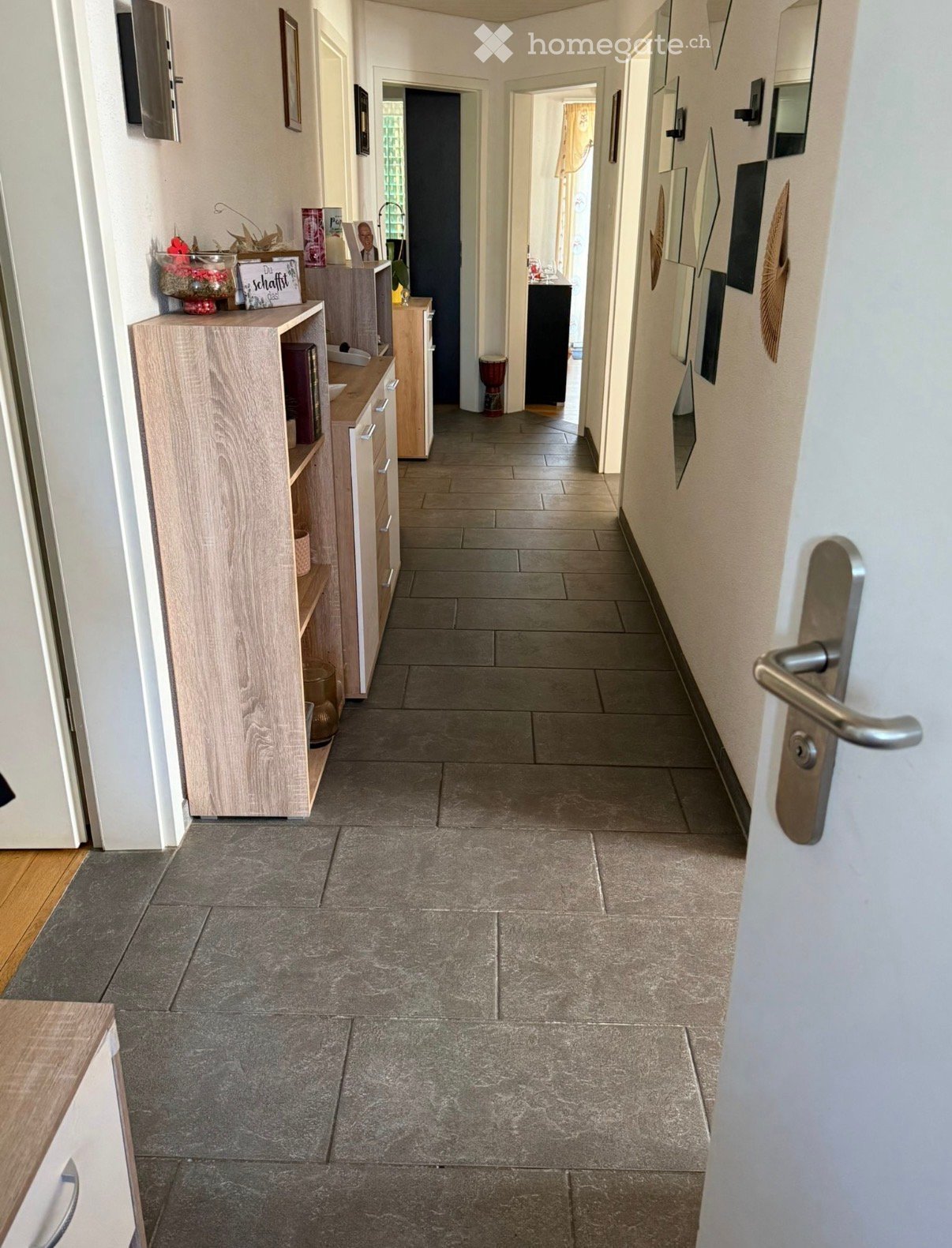 A clean, tiled hallway with light-colored walls, wooden cabinetry on the left, and several doors leading to other rooms.