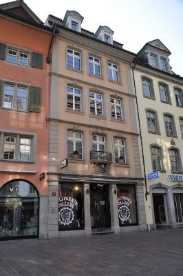 Three-story urban building with classic European architecture, featuring large windows, decorative balconies, and storefronts at ground level.