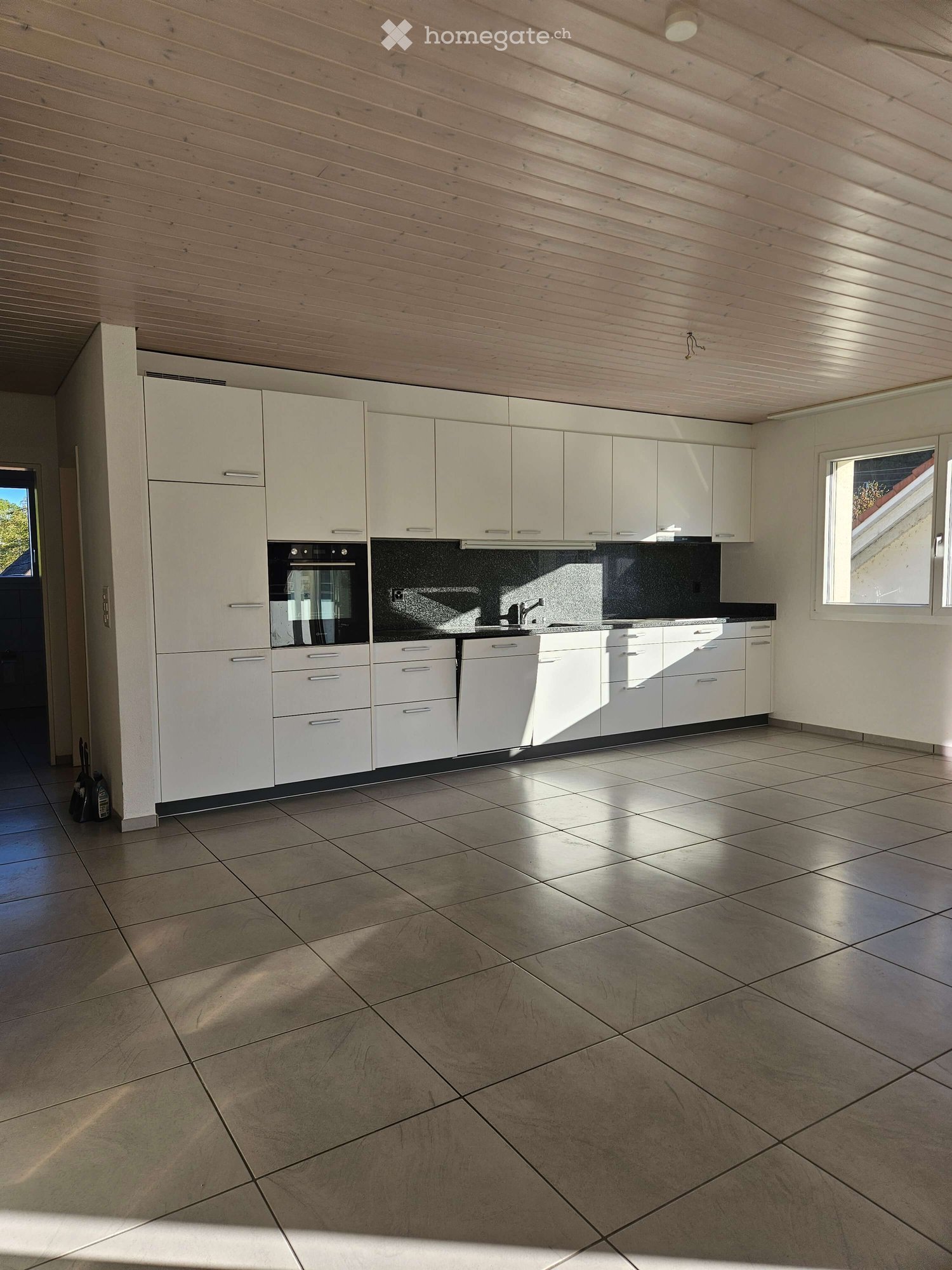 Empty kitchen with white cabinets, stainless steel appliances, and tiled flooring. Natural light streams in from a window on the right side.