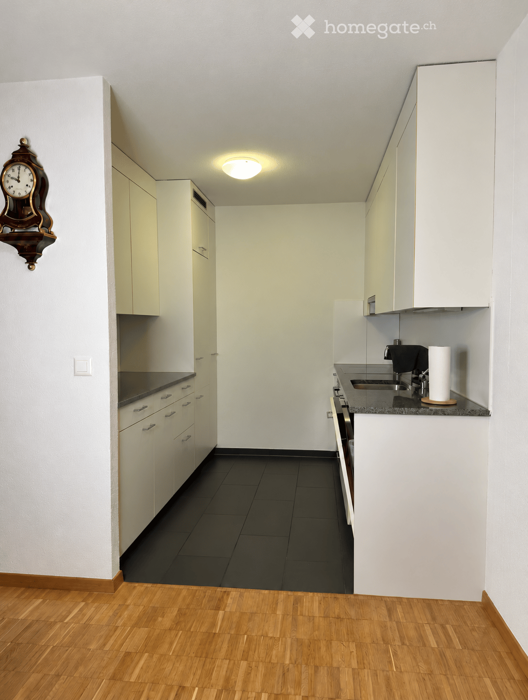 Modern kitchen with white cabinets, black countertops, and a dark tiled backsplash. Features a sink, faucet, oven, and a wall-mounted clock.