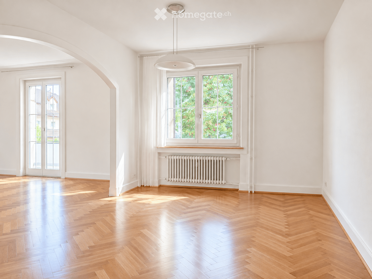 Empty room with wooden herringbone flooring, white walls, large windows with white frames, and a modern pendant light fixture.