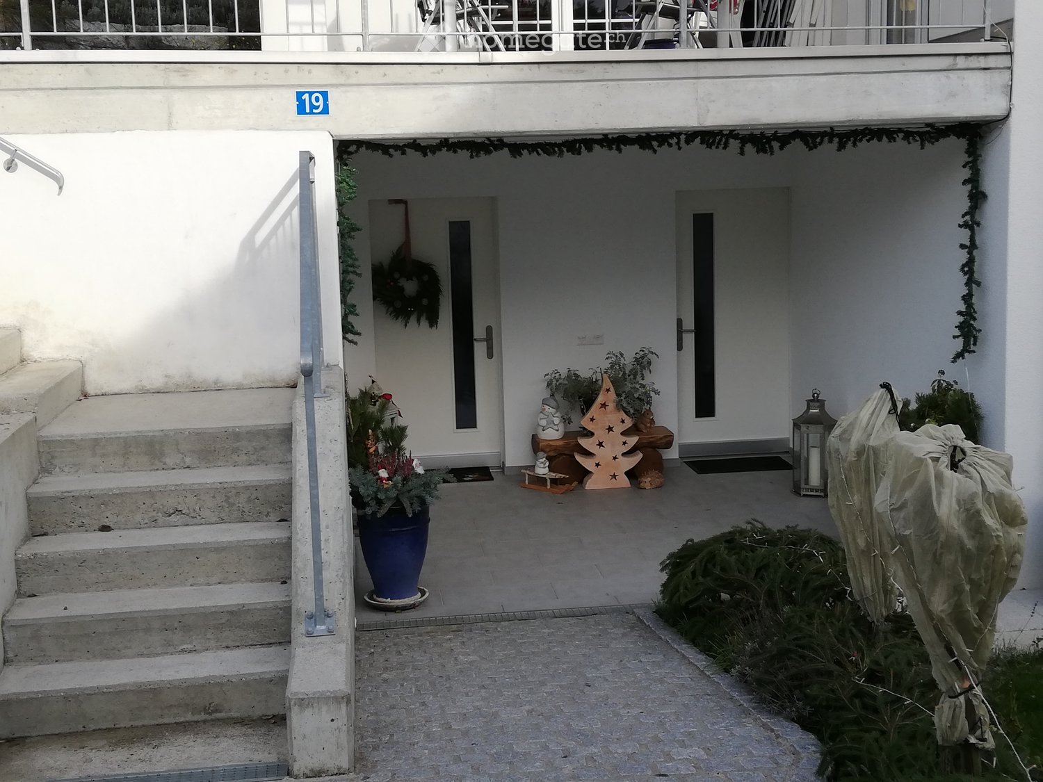 Entrance area with steps leading up to a door, decorated with a Christmas wreath, potted plants, and a small wooden Christmas tree ornament.