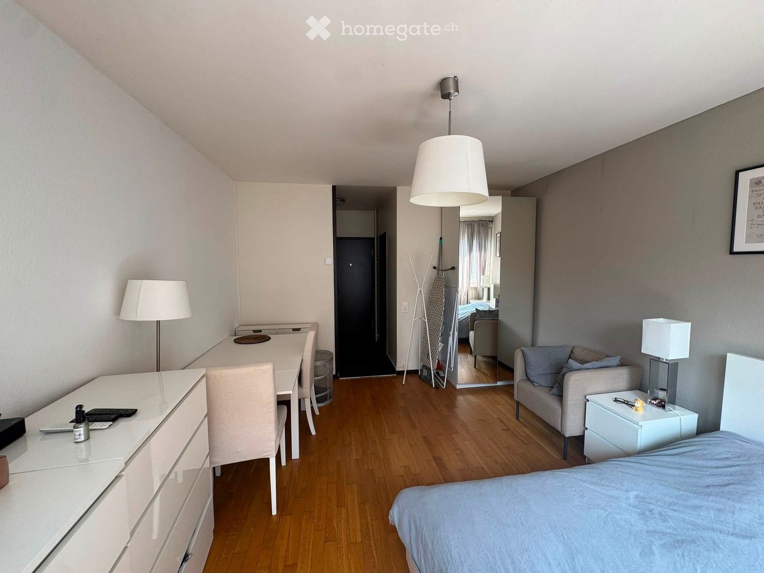 A modern, well-lit room with a bed, sofa, dining table, chairs, and a dresser. The room features wooden flooring and white walls.