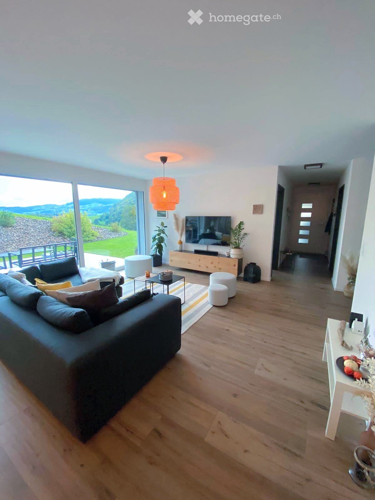 Modern living room with large windows offering a scenic view, featuring a dark grey sectional sofa, coffee table, TV, and decorative plants.