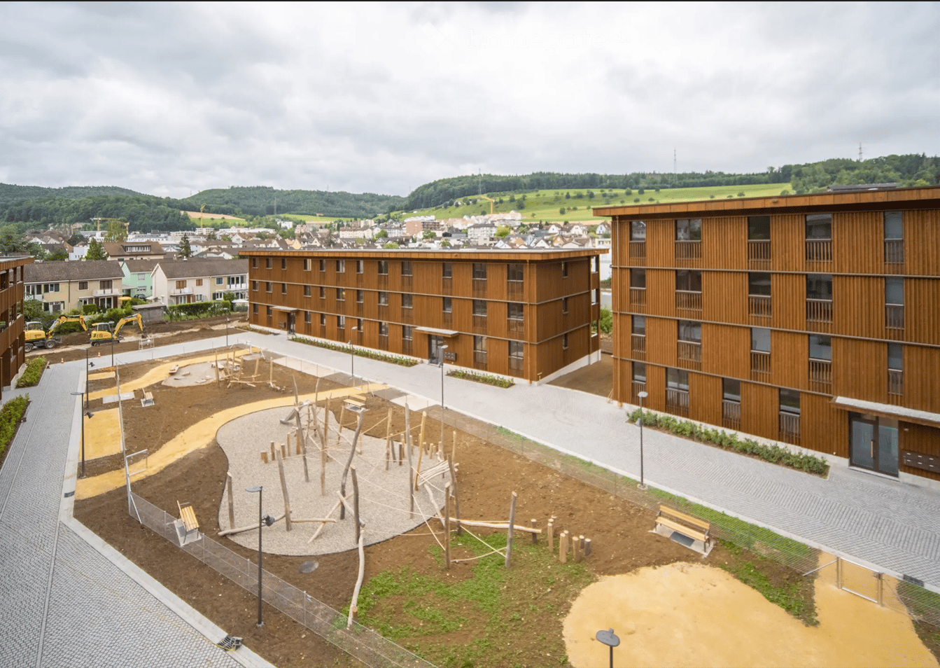 Modern multi-story residential building with wooden facade, surrounded by playground equipment and paved walkways.
