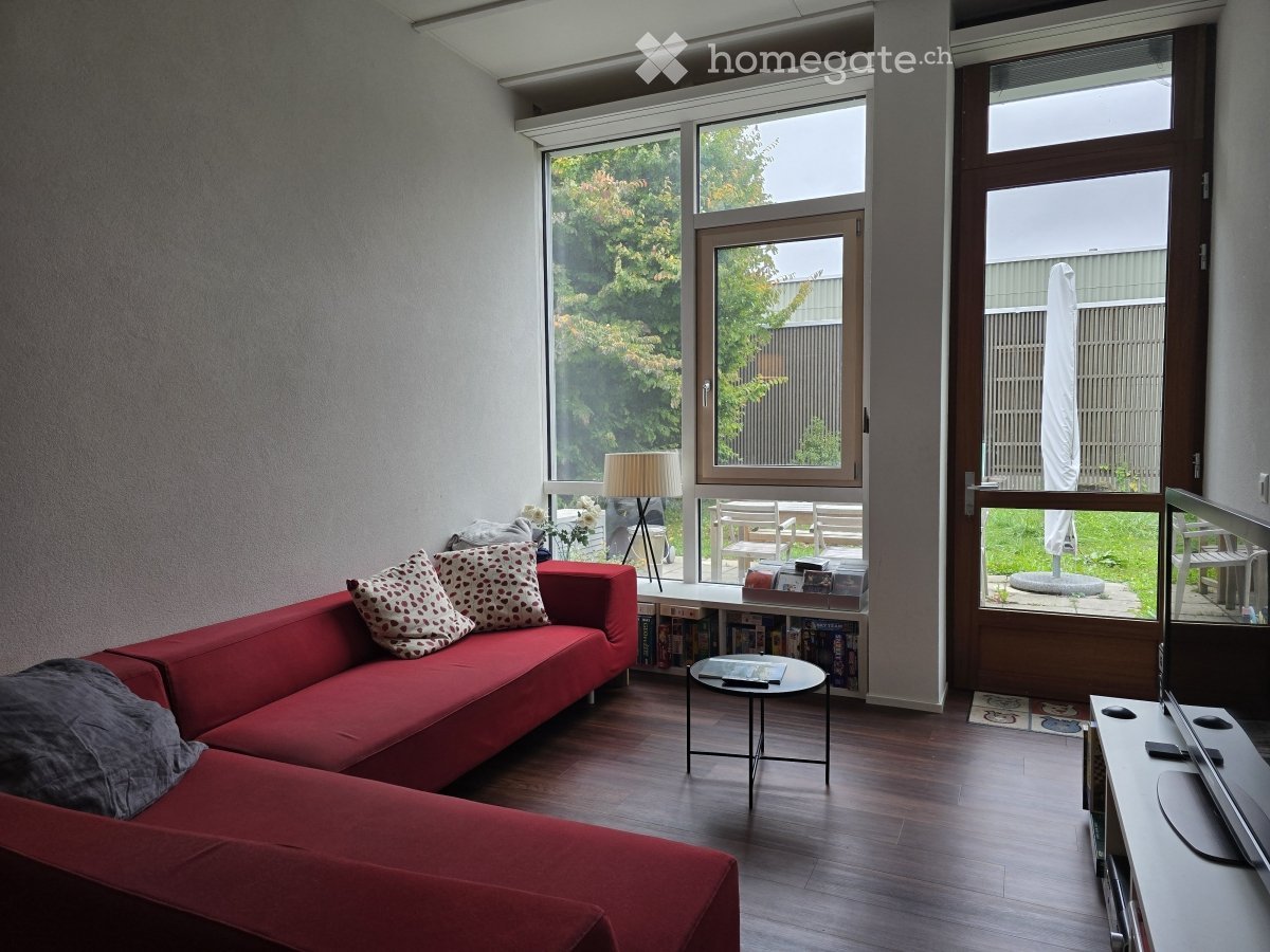 Modern living room with large windows, red sectional sofa, coffee table, bookshelf, and TV stand.