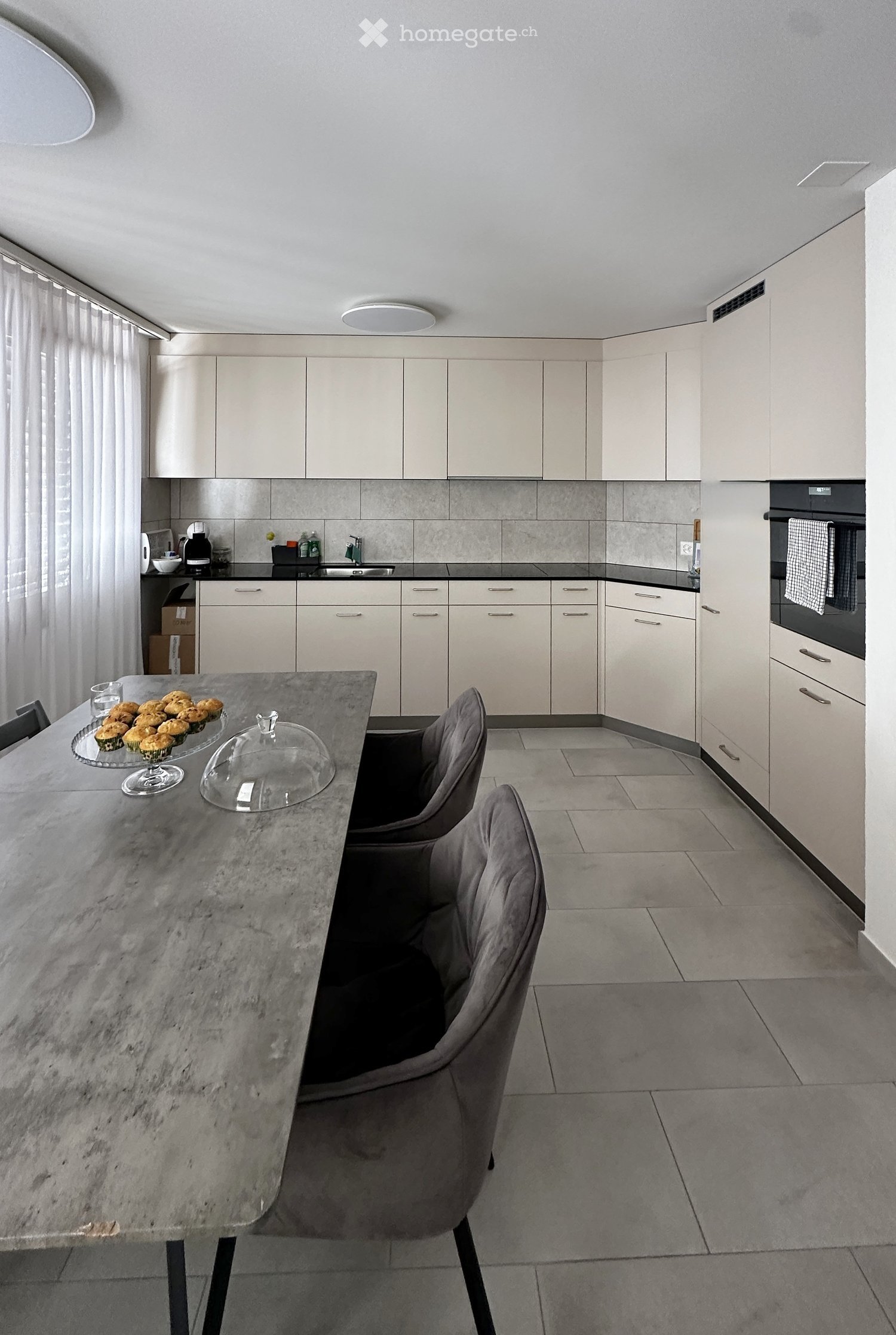 Modern kitchen with white cabinets, grey tiled backsplash, and a central island with a marble countertop. The island features a glass dome and a plate of muffins, with two grey chairs around it. A sink, oven, and various small appliances are visible along