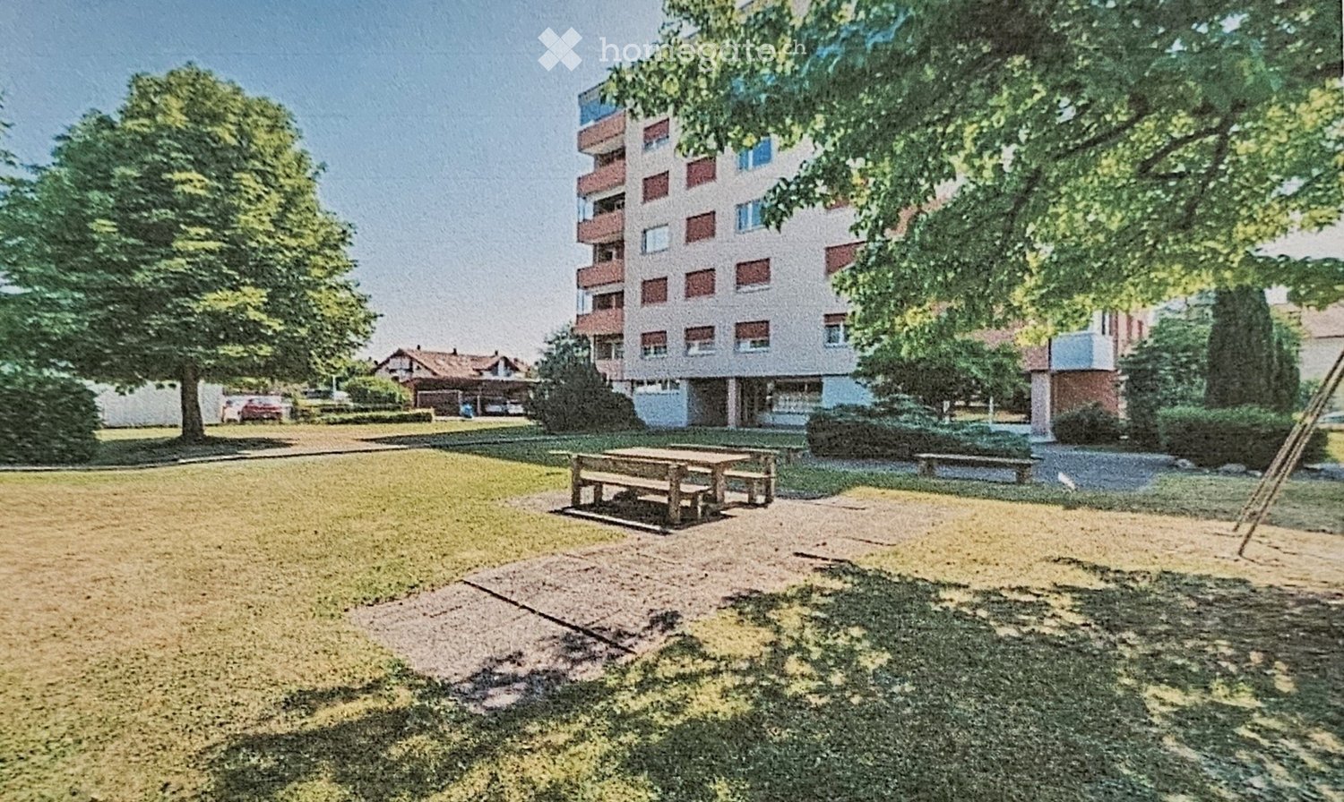 A grassy park area with a wooden picnic table, benches, trees, and a multi-story residential building in the background.