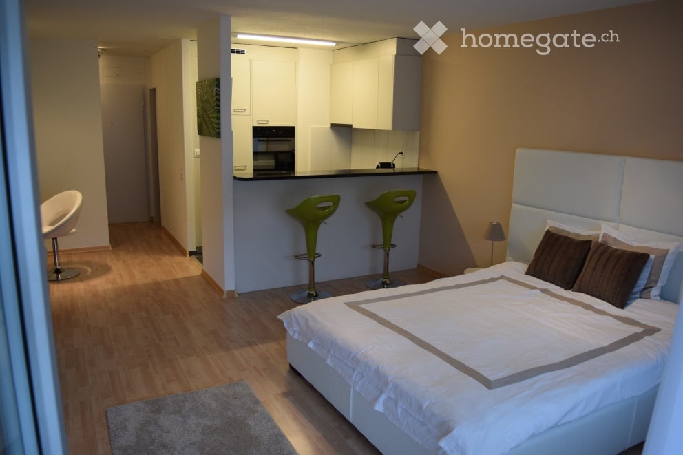 A modern, well-lit bedroom with a large bed, white headboard, and brown pillows. A small kitchen area is visible with white cabinets, a countertop, and two green bar stools. A white chair is placed near the entrance, and a small lamp sits on a bedside tab
