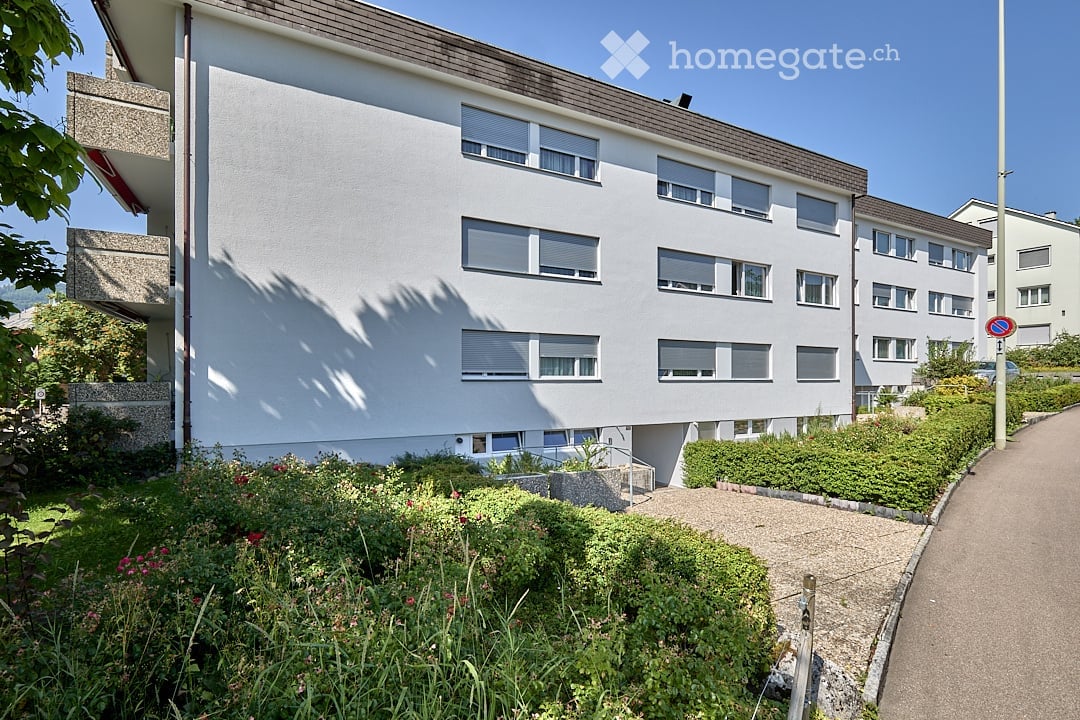 Modern multi-story residential building with white facade, multiple windows, and a landscaped garden area in front.