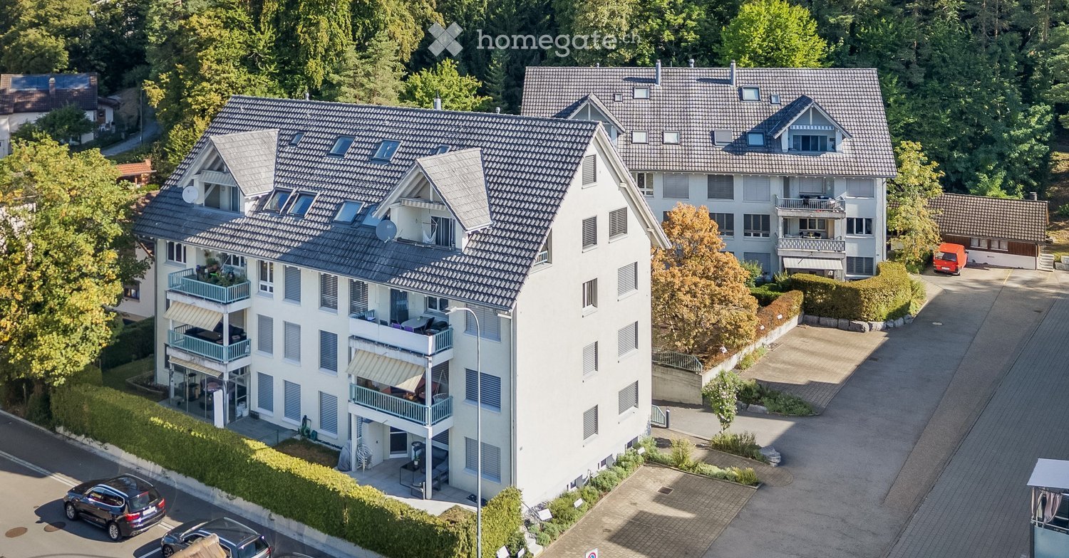 Aerial view of a multi-story residential building with multiple balconies, surrounded by greenery and parking spaces.