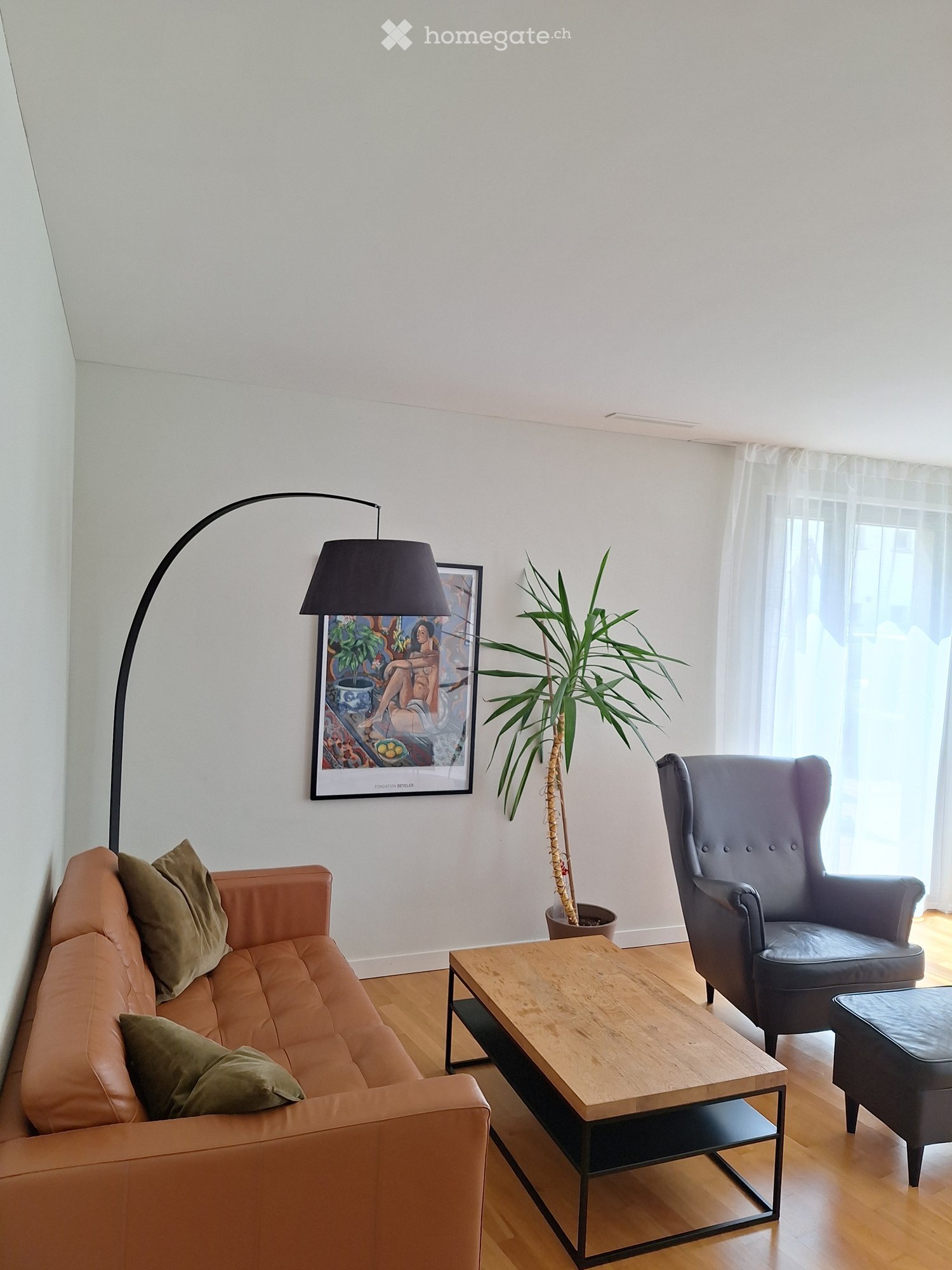 Modern living room with a light brown leather sofa, wooden coffee table, grey armchair, floor lamp, potted plant, and artwork on the wall.