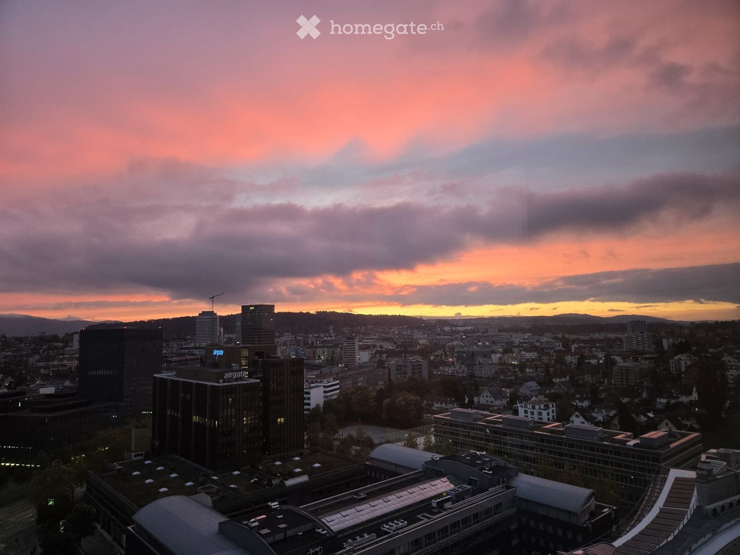 Aerial view of a city with multiple high-rise buildings, a sunset sky, and a distant mountain range.