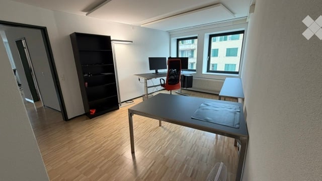 Bright, modern office space with wooden flooring, large windows, a desk with a computer, a bookshelf, and a comfortable chair.