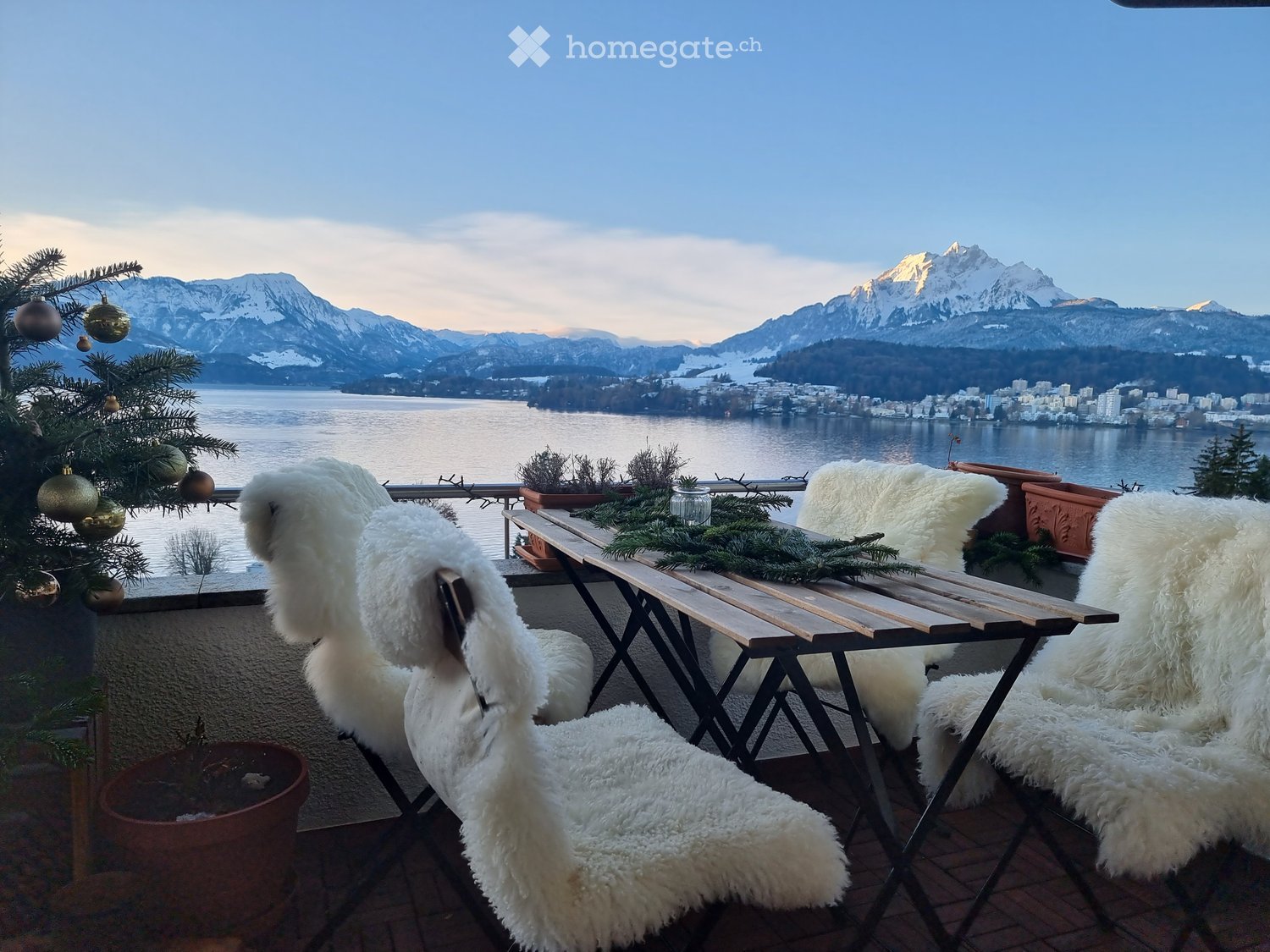 Balcony with a scenic mountain and lake view, decorated with a Christmas tree, a wooden table, and white fur-covered chairs.