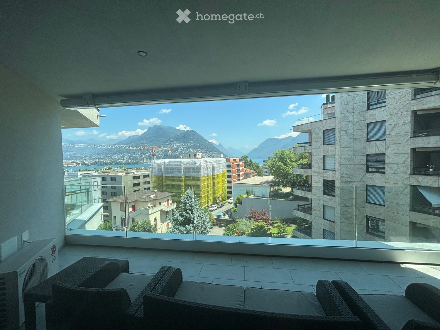Balcony with large glass windows, offering a scenic view of mountains and a lake, furnished with a sofa and chairs.