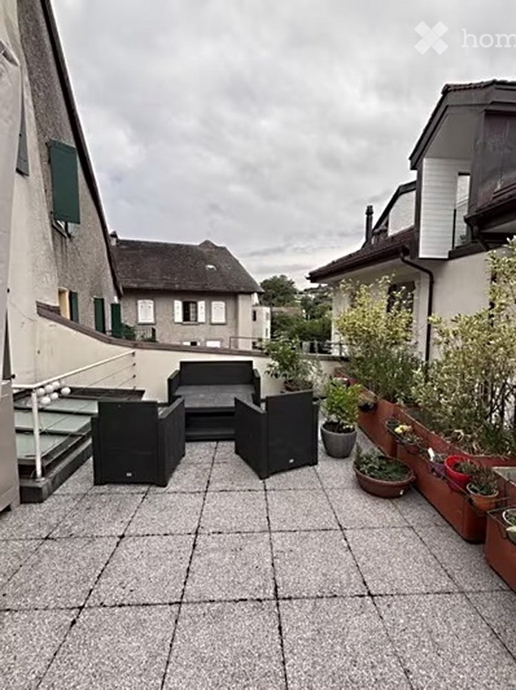 A modern outdoor patio with a grey tiled floor, black seating area including a sofa and chairs, and several potted plants arranged along the edge.