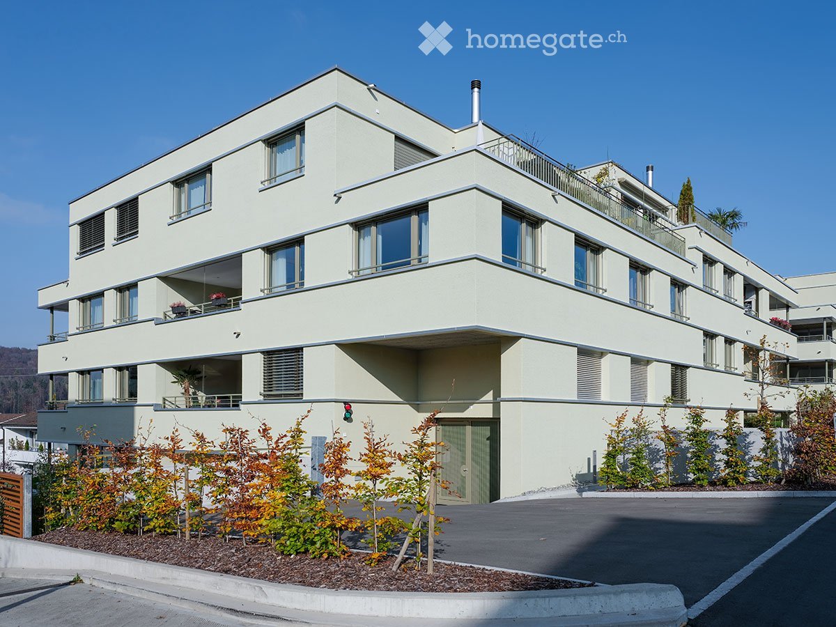 Modern three-story residential building with light beige facade, multiple balconies, and small garden area in front.
