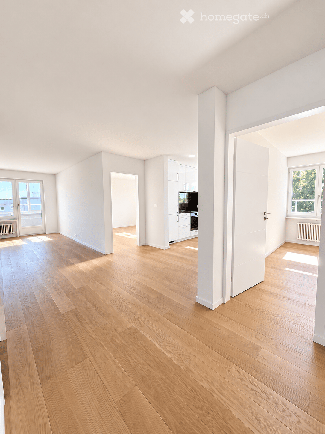 Bright, empty room with light wooden flooring, white walls, and large windows allowing natural light. The space features a central column and an open doorway leading to another area with white cabinetry and appliances.