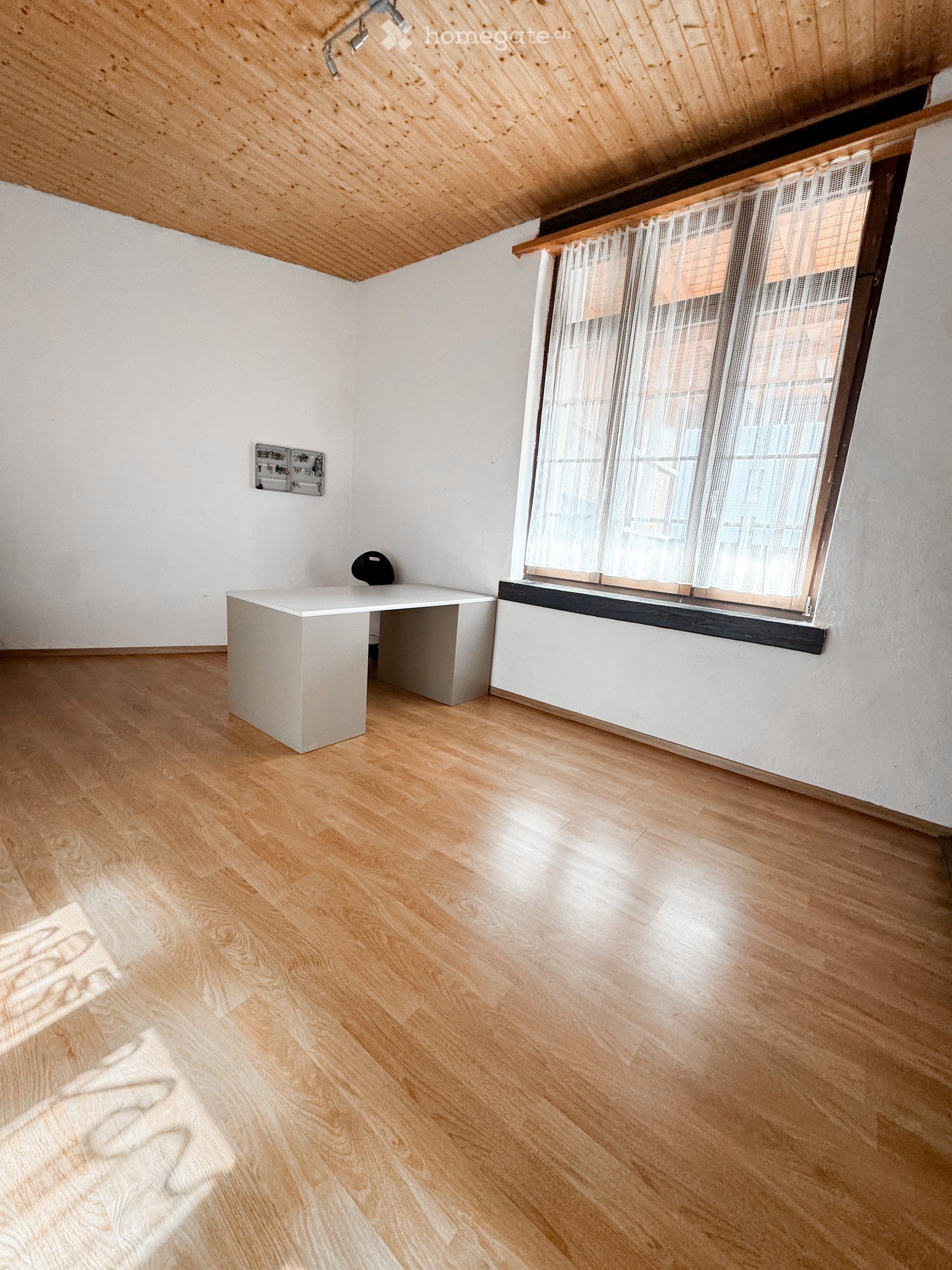 Empty room with wooden flooring, white walls, a large window with sheer curtains, a small white table, and a black chair.