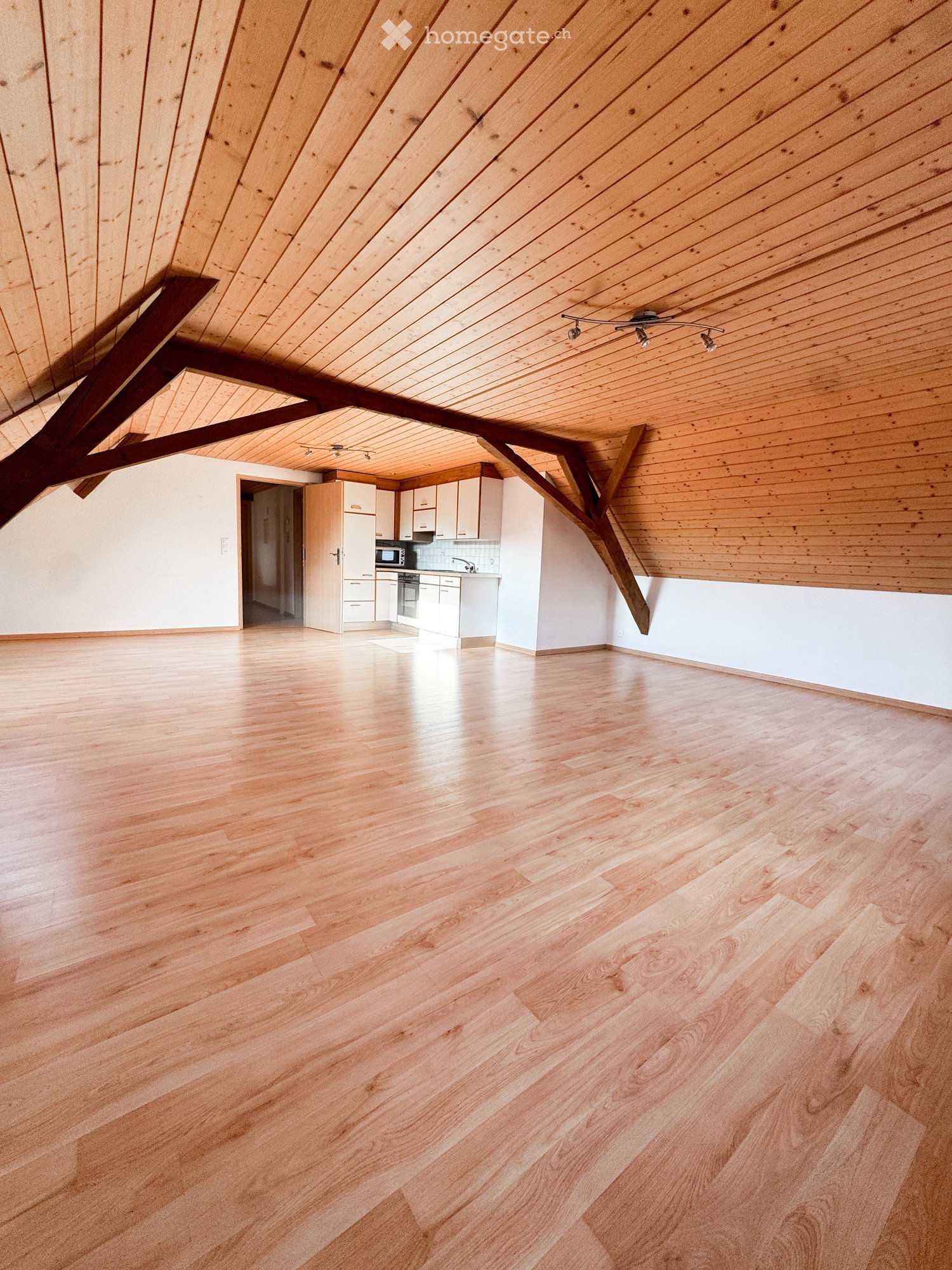 Empty room with wooden flooring and white walls, featuring exposed wooden beams on the ceiling and a small kitchen area visible in the background.