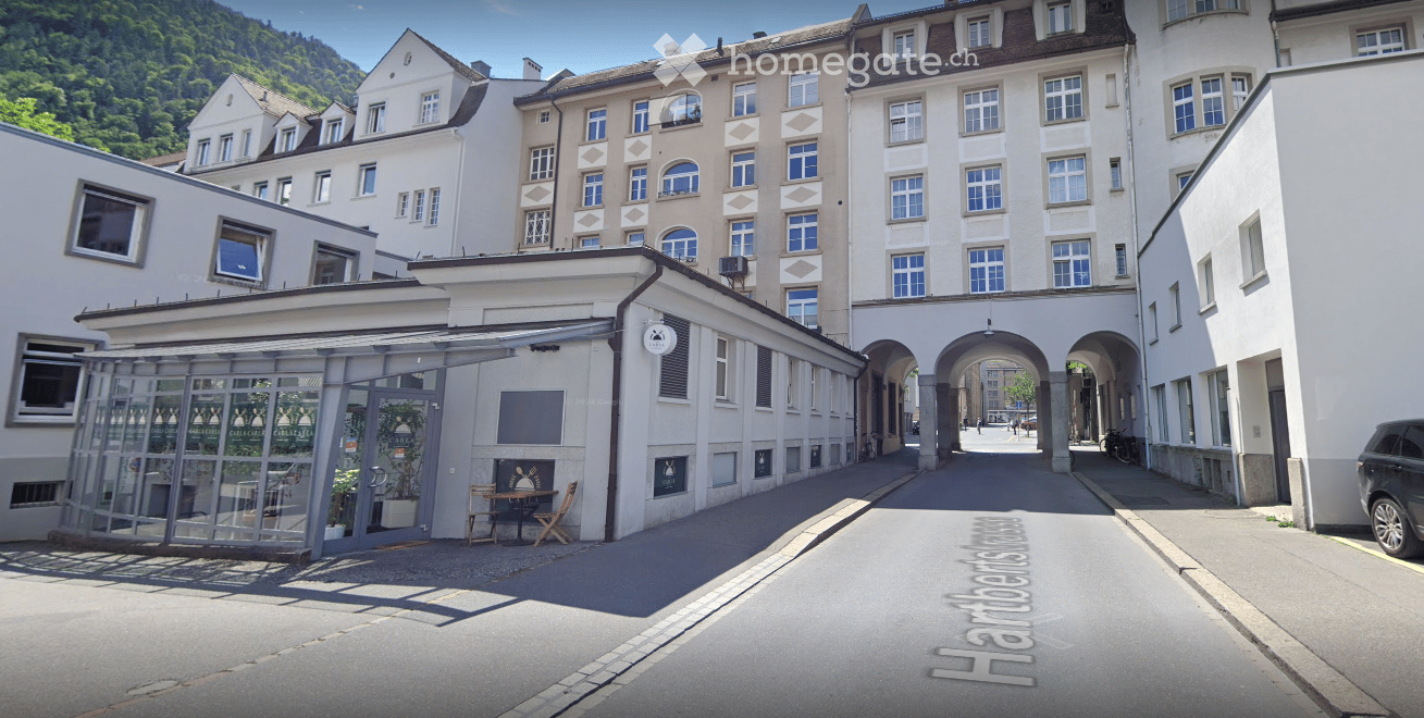 A row of multi-story buildings with classic European architecture, featuring large windows, arched entrances, and a small outdoor seating area with tables and chairs.