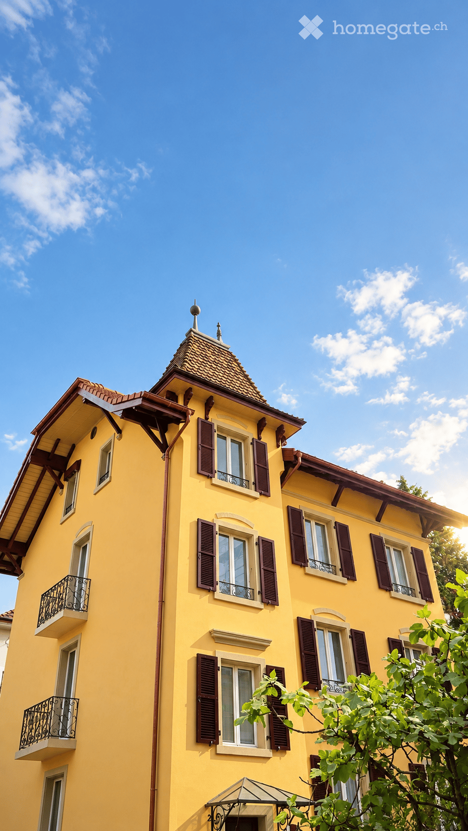 A multi-story yellow house with a red-tiled roof, multiple windows with dark shutters, and small balconies with black wrought-iron railings.