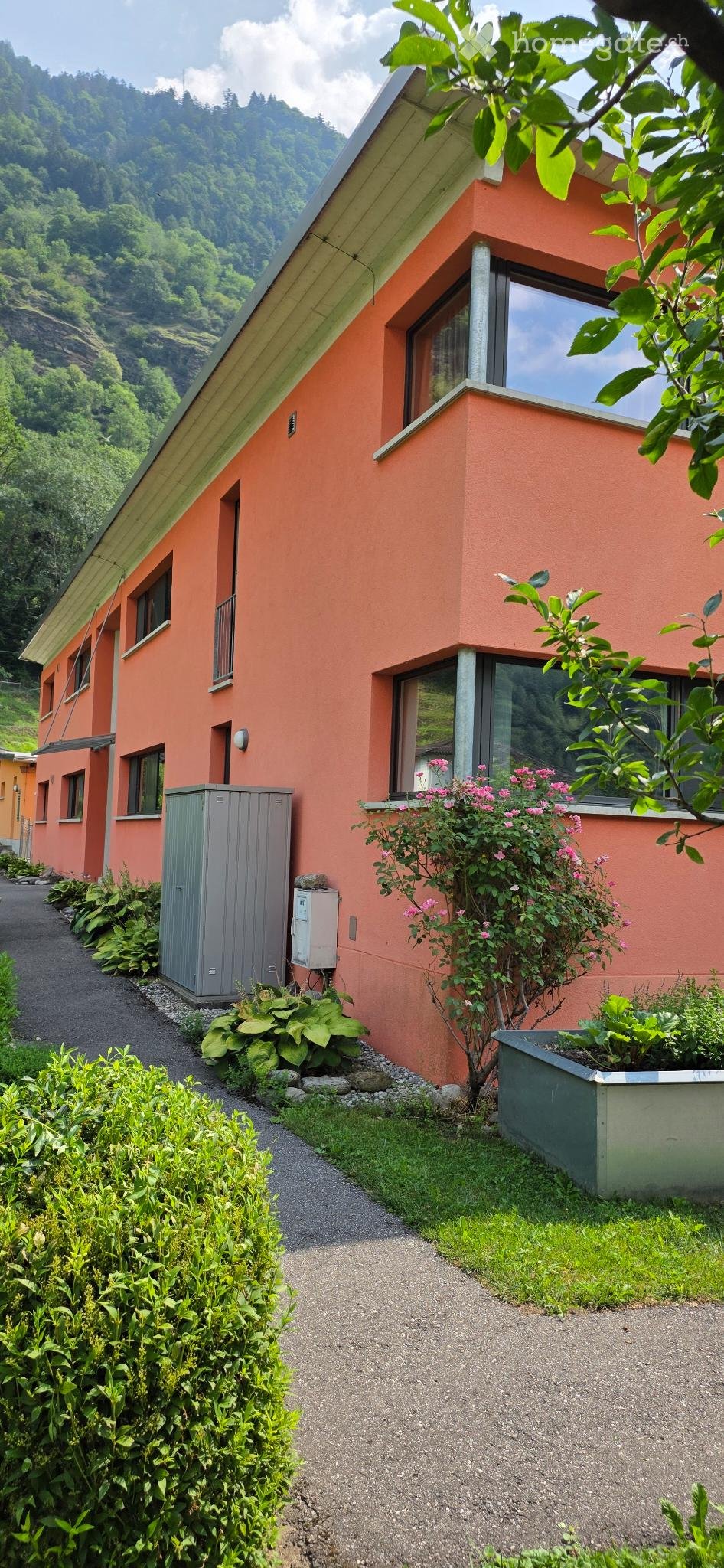 A two-story orange house with large windows, surrounded by greenery and a gravel pathway.