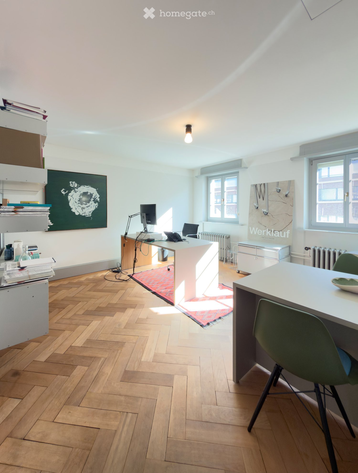 Bright, modern office space with wooden herringbone flooring, white walls, and large windows. Features a desk with a computer, a chair, a shelf with books and art, and a small table with a green chair.