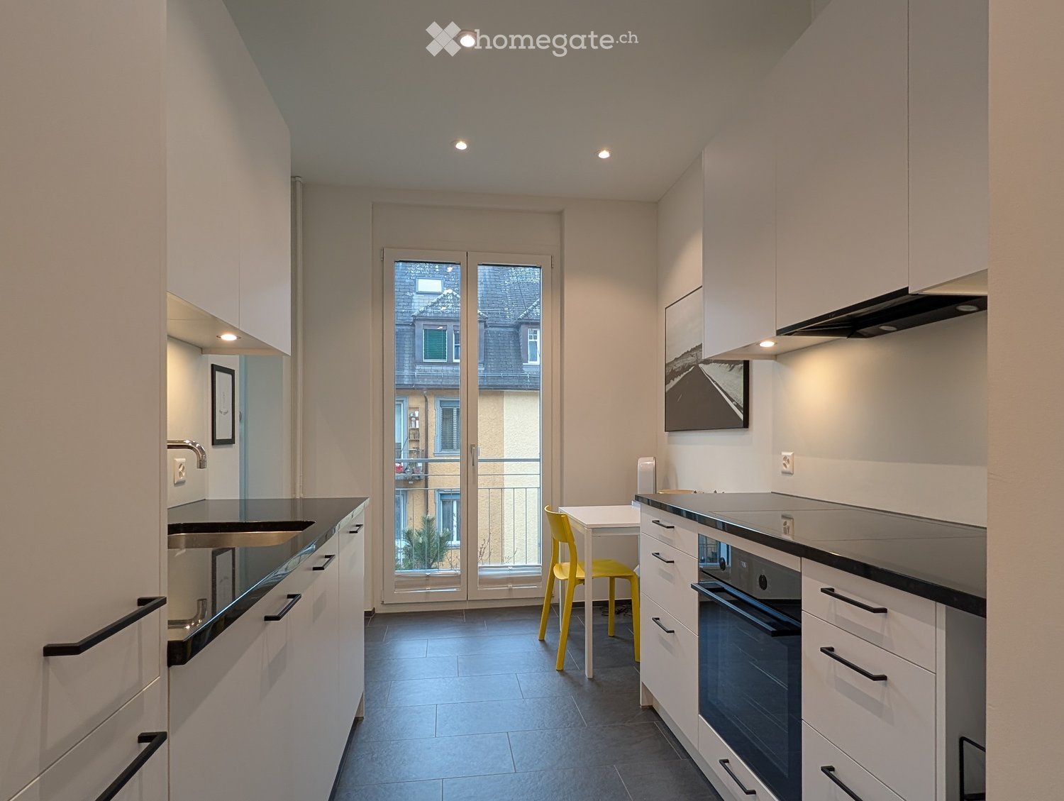 Modern kitchen with white cabinets, black countertops, a sink, an oven, and a small yellow chair and table near the sliding glass door leading to a balcony.