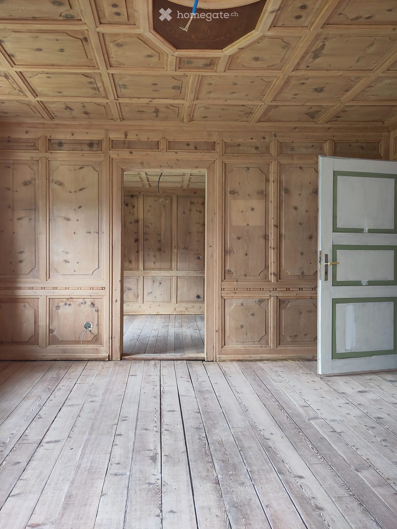 Empty room with wooden walls, wooden ceiling, and wooden floor. A white door with green trim is visible on the right side.