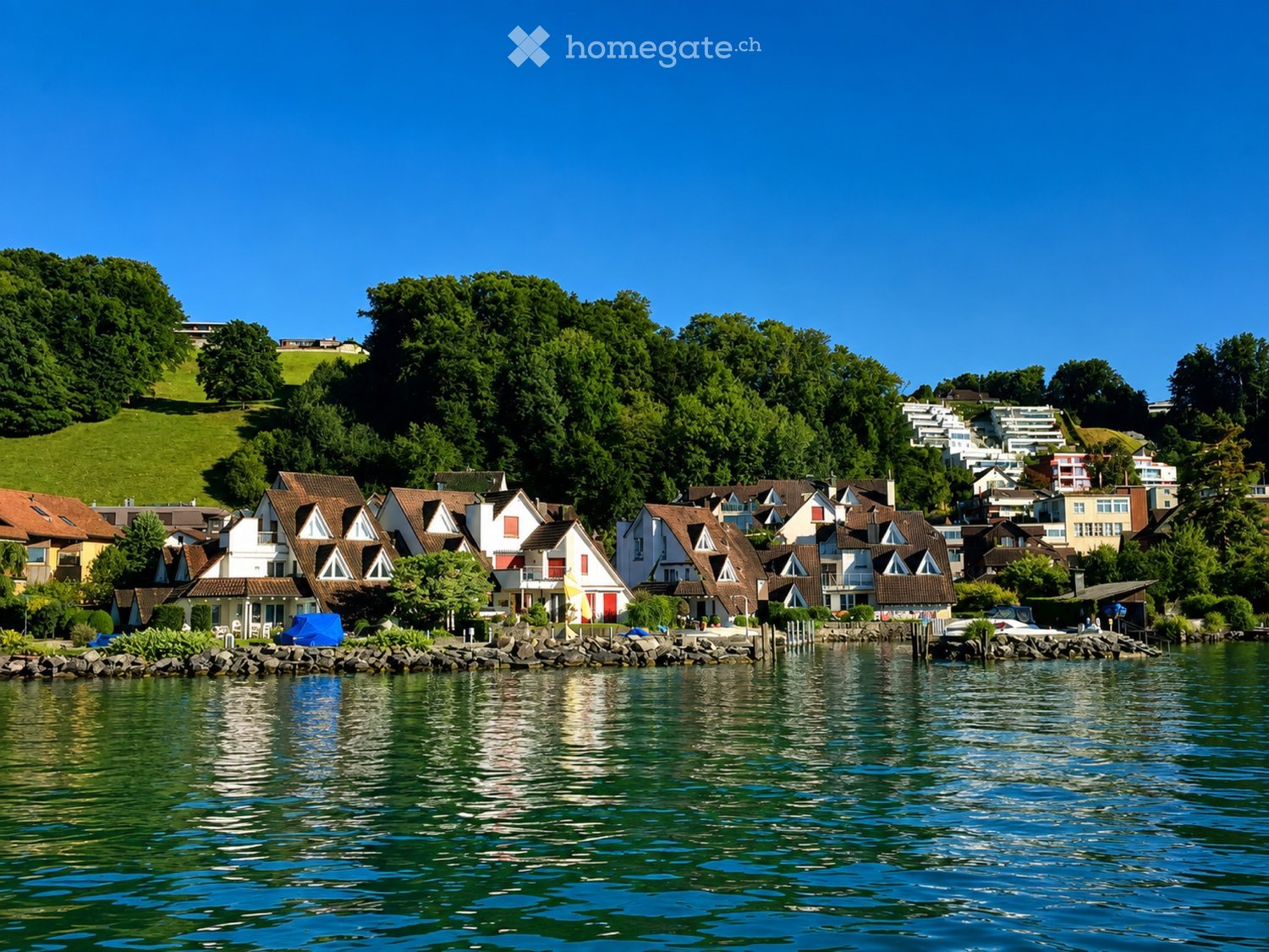 Residential area with houses along a lakeshore, surrounded by lush green trees and hills.