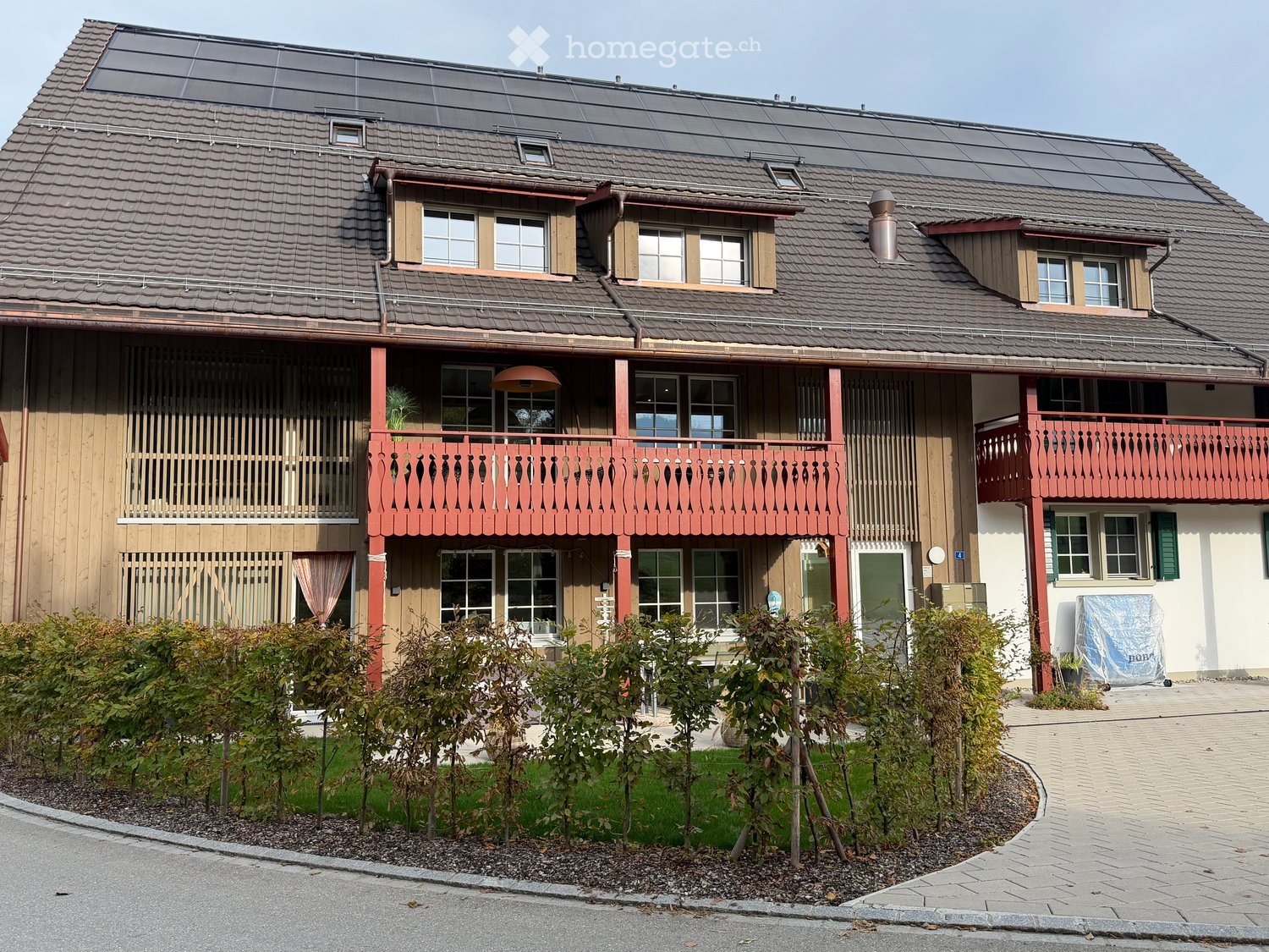 Two-story house with red wooden balconies, multiple windows, solar panels on the roof, and a well-maintained garden with small trees and shrubs.