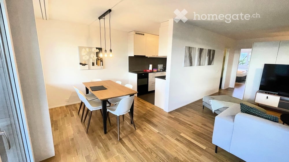 Modern, open-plan living and dining area with a white sofa, coffee table, dining table with chairs, and a compact kitchen featuring cabinets, an oven, and a fridge. The space is well-lit with pendant lights and has a sleek, contemporary design.