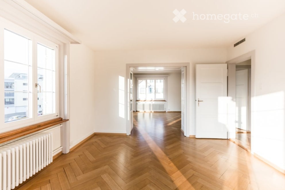 Empty room with wooden herringbone flooring, white walls, large windows, and a radiator. No furniture or equipment present.