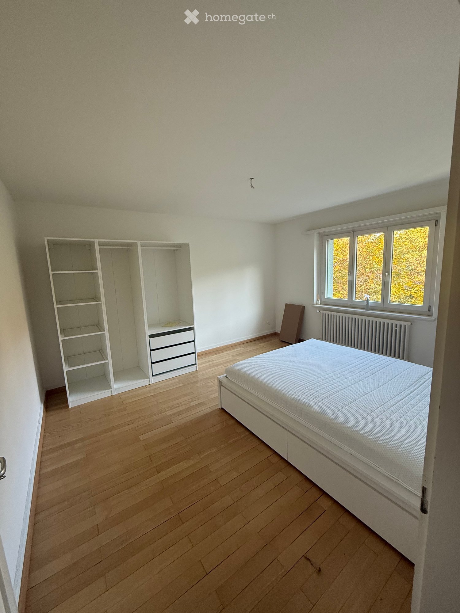 Minimalist bedroom with wooden flooring, white walls, a single bed, a built-in wardrobe, and a radiator under the window.