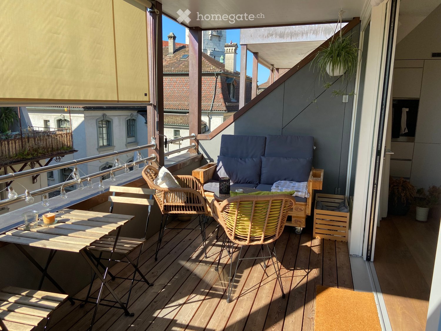 Balcony with wooden decking, sofa, table, chairs, and hanging plants.