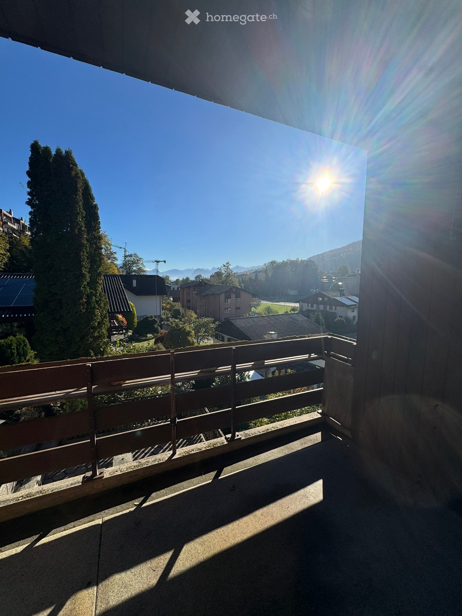 Balcony with wooden railing, overlooking a scenic town with houses, trees, and mountains under a clear blue sky.