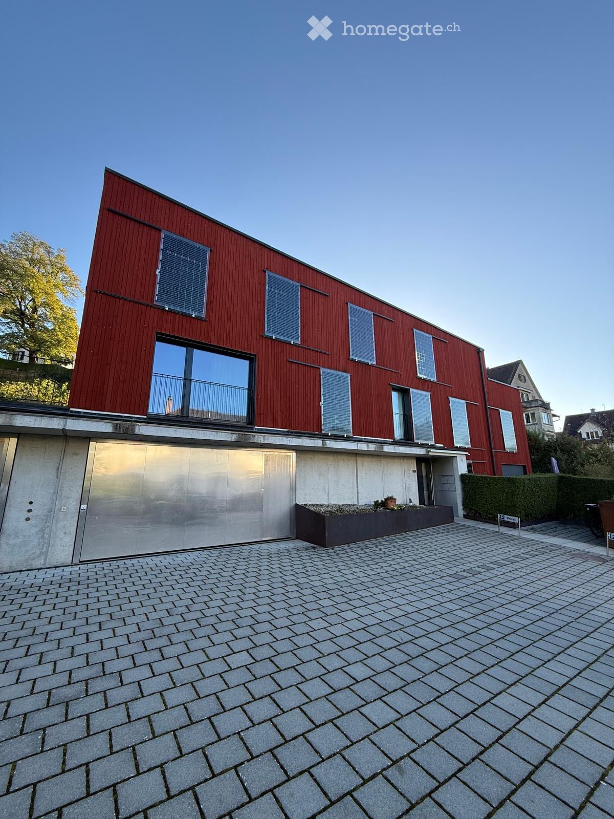 Modern two-story house with red wooden facade, large windows, and a concrete lower level. Features include a raised ground floor, a small garden area, and a paved driveway.