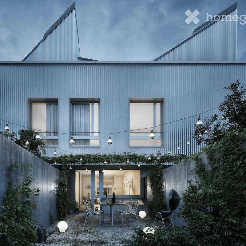 Modern two-story house with grey exterior, large windows, and a small outdoor seating area with a table and chairs surrounded by greenery and string lights.