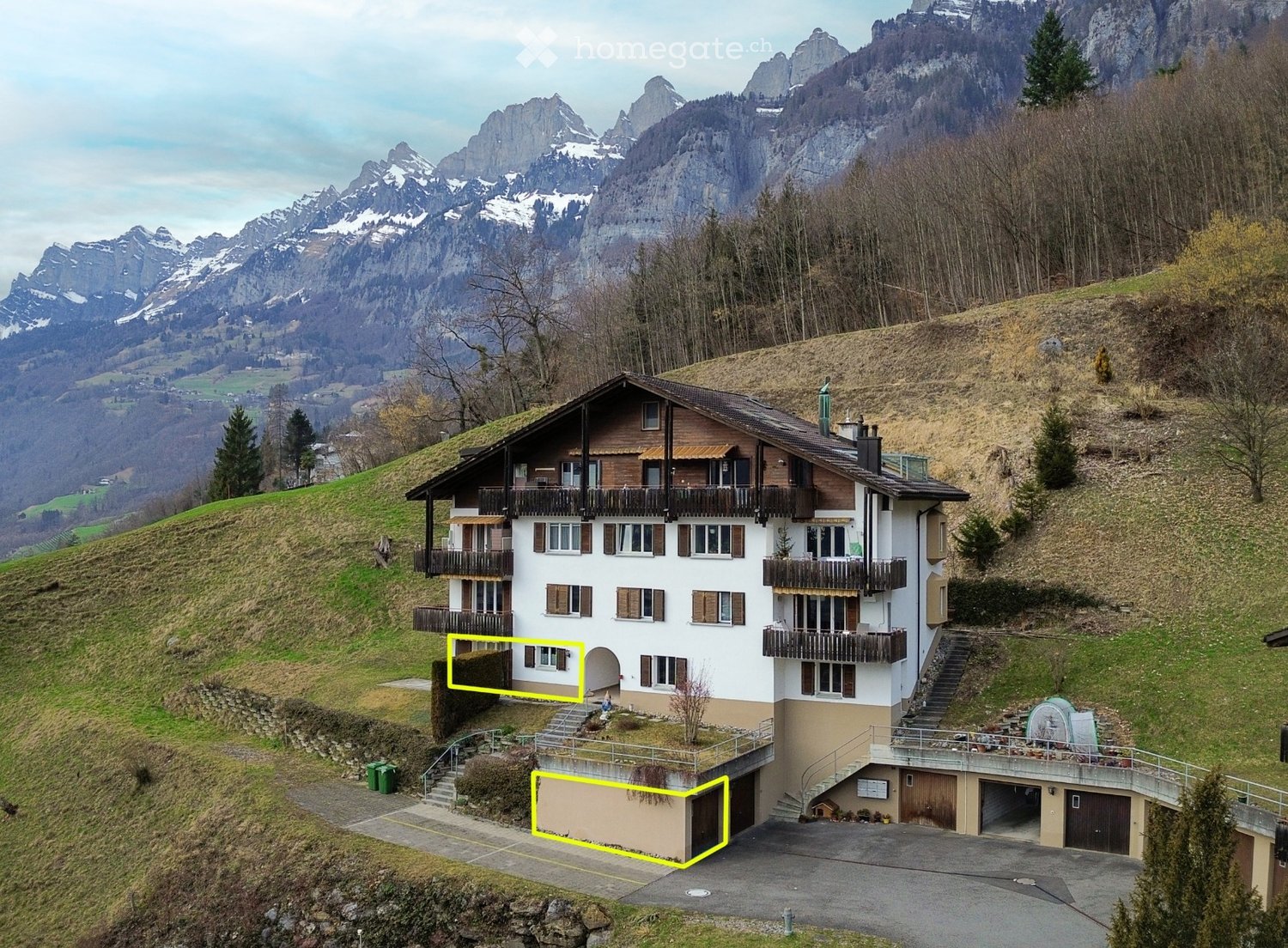 A multi-story house with balconies, situated on a hillside with a scenic mountain view in the background. The house features a garage and is surrounded by greenery.